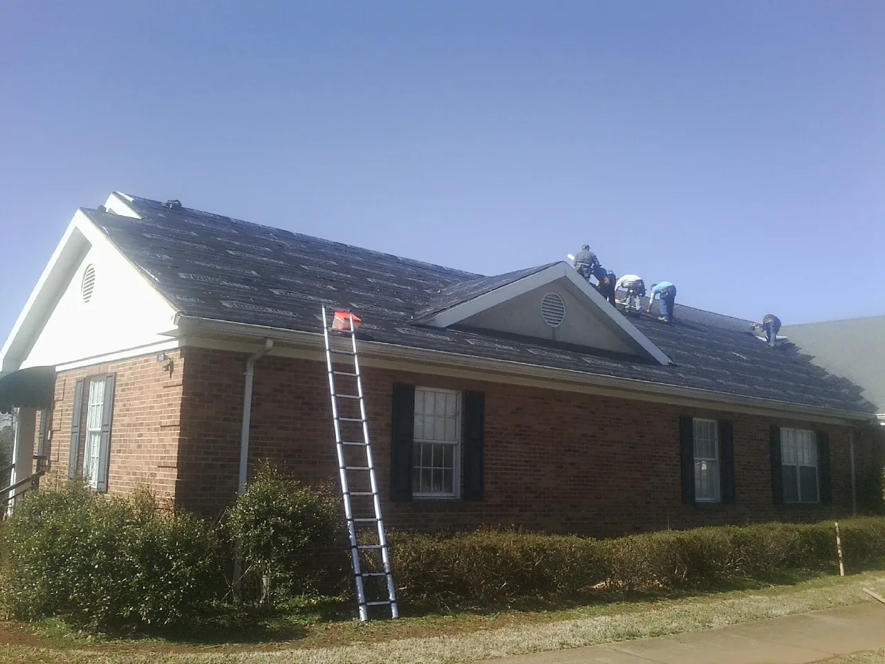 Crew tearing shingles off a brick ranch roof with a ladder set — Emergency Roof Repair work by our crew in Lagrangeville