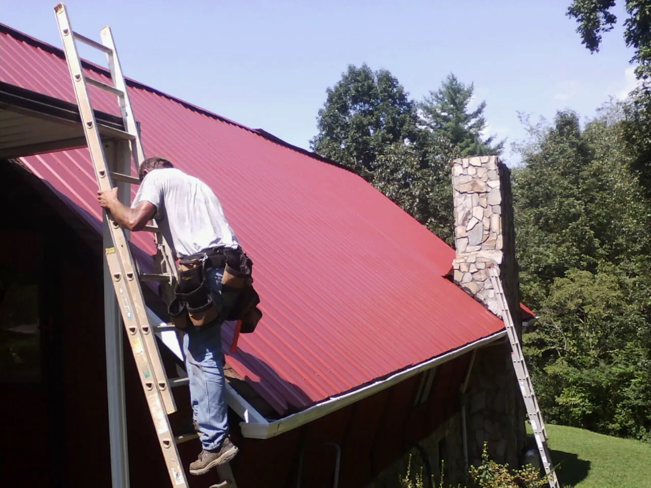 Roofer on a ladder beside a red metal roof and stone chimney — Soffit & Fascia Repair by Roofing Brothers in Lagrangeville