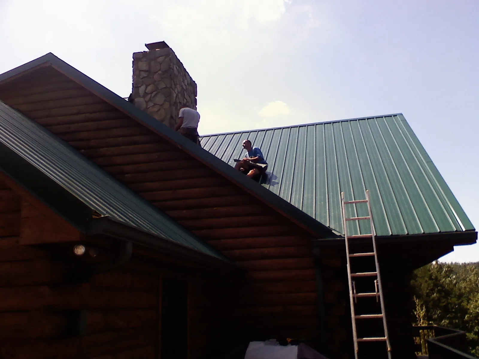 Roofers installing green metal panels beside a stone chimney on a log home in Lagrangeville
